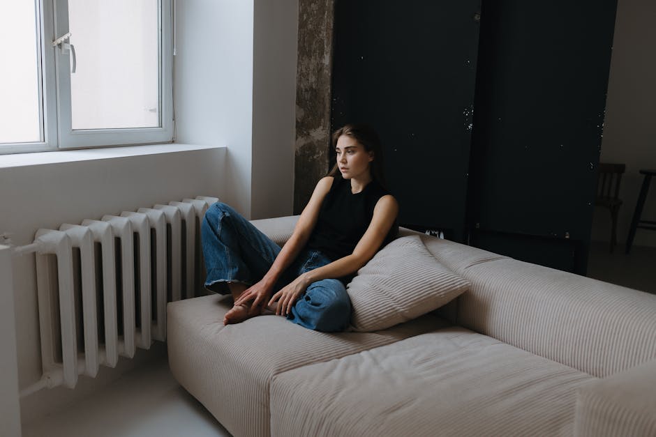 A serene young woman sits barefoot on a sofa in a minimalist studio.