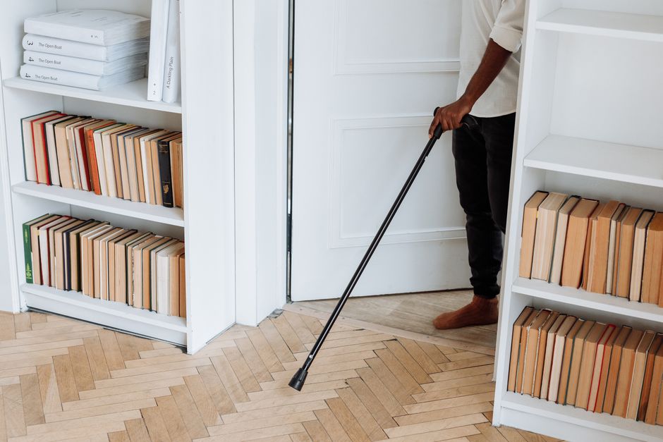 A person using a walking cane to move around a room filled with bookshelves indoors.