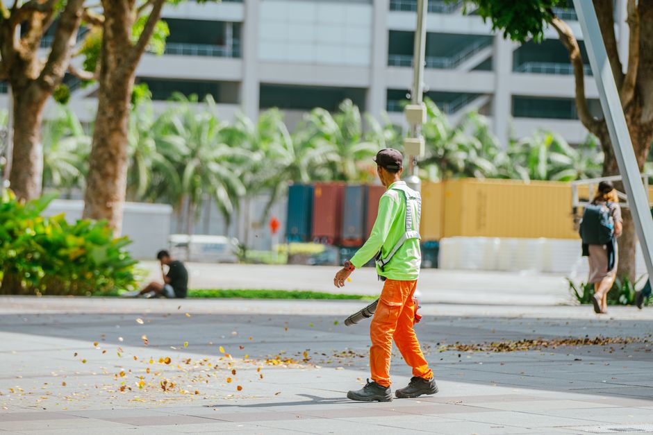 Street cleaner using a leaf blower in an urban park setting on a sunny day.