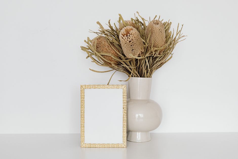 A ceramic vase with dry banksia flowers next to a woven picture frame on a white background.